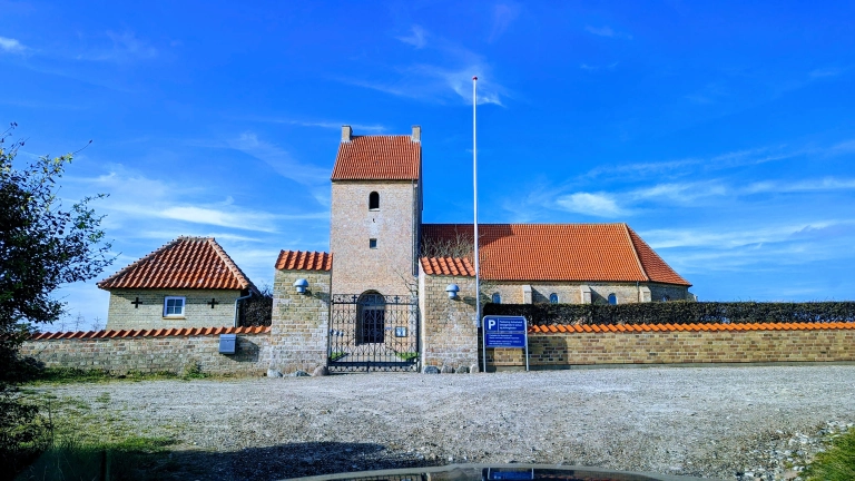 Eine Steinkirche mit zentralem Turm und rotem Ziegeldach steht hinter einem Zaun, davor ein Kiesparkplatz unter strahlend blauem Himmel.