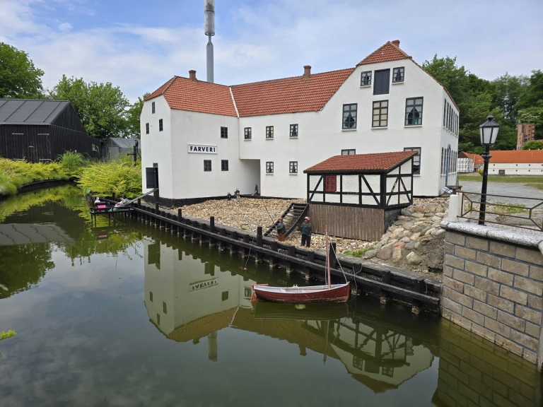 Ein weißes Gebäude mit roten Dächern spiegelt sich in einem Kanal, mit einem kleinen Steg, einem Boot und mehreren menschlichen Miniaturfiguren am Wasser.