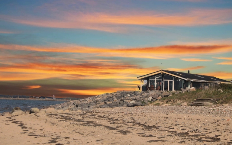 Ein kleines Haus steht an einer felsigen Küste bei Sonnenuntergang, mit bunten Wolken am Himmel und Sand im Vordergrund.